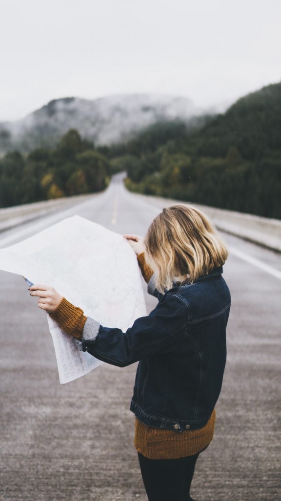 A woman looking at map while standing in a road leading off to the mountains on the horizon beyond her.  A visual analogy of the Needs Analysis Process starting point.